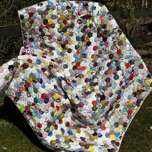 May include: A handmade quilt with a white background and a pattern of colorful, hexagonal fabric patches. The patches are in various colors and patterns, including floral, striped, and solid fabrics. The quilt is spread out on green grass.