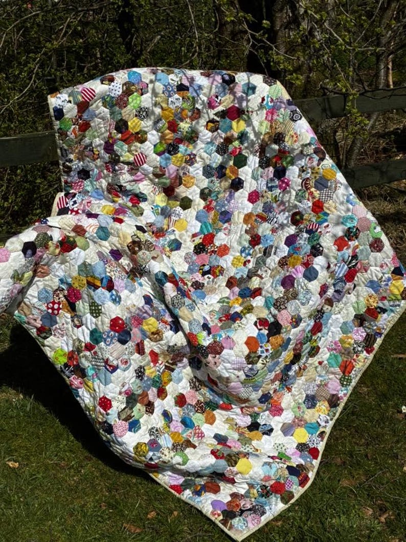 May include: A handmade quilt with a white background and a pattern of colorful, hexagonal fabric patches. The patches are in various colors and patterns, including floral, striped, and solid fabrics. The quilt is spread out on green grass.