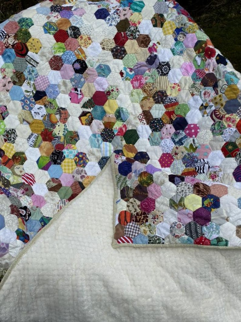 May include: A handmade quilt featuring a patchwork design of colorful hexagonal fabric pieces. The quilt's top is a vibrant mix of patterns and colors, while the underside is a soft, cream-colored material.