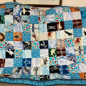 May include: A blue and white patchwork quilt featuring a variety of airplane and sky-themed fabric squares. The quilt is edged with a blue border and has a small airplane design in the centre.