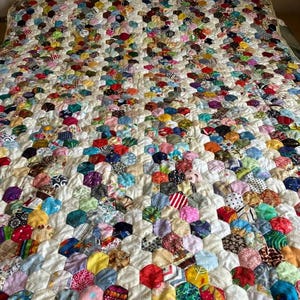 May include: A handmade quilt with a colorful patchwork design. The quilt features a pattern of hexagonal fabric pieces in various colors and patterns, including floral, striped, and solid fabrics. The quilt is on a bed with a metal headboard.