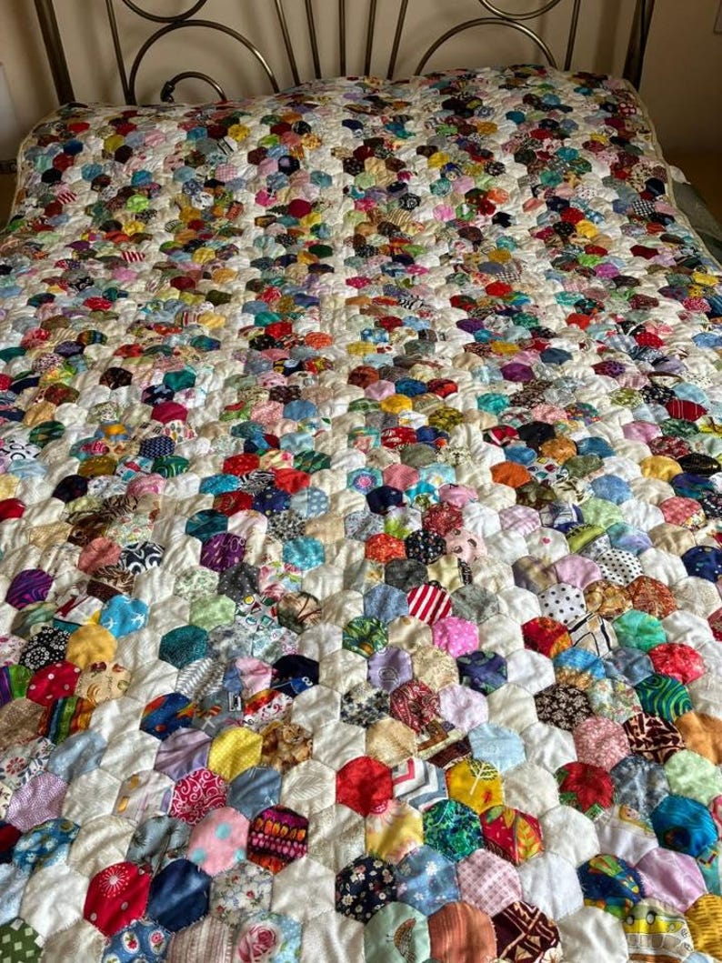 May include: A handmade quilt with a colorful patchwork design. The quilt features a pattern of hexagonal fabric pieces in various colors and patterns, including floral, striped, and solid fabrics. The quilt is on a bed with a metal headboard.