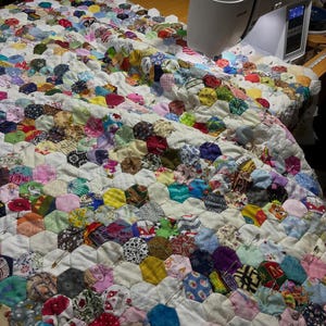 May include: A colorful handmade quilt in progress, featuring a patchwork design of hexagonal fabric pieces in various patterns and colors. A white sewing machine is positioned on the right side of the quilt, with the needle poised to stitch the fabric together.
