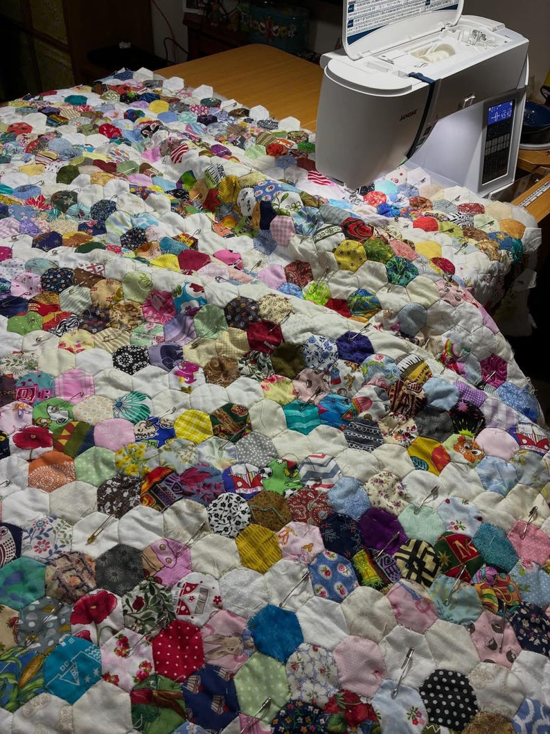 May include: A colorful handmade quilt in progress, featuring a patchwork design of hexagonal fabric pieces in various patterns and colors. A white sewing machine is positioned on the right side of the quilt, with the needle poised to stitch the fabric together.