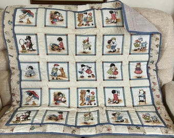 May include: A cream-colored quilt featuring a repeating pattern of children in various activities, framed in blue squares. The quilt has a blue border and a patterned edge with small illustrations. The overall design is a vintage, storybook theme.