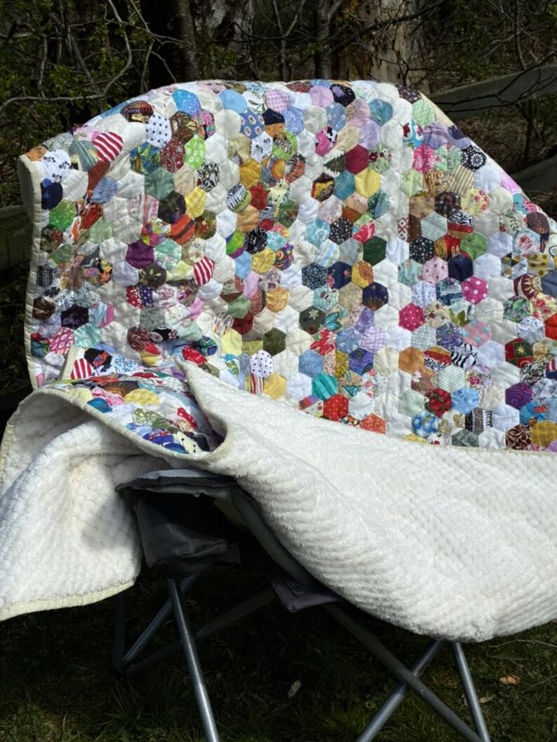 May include: A colorful handmade quilt with a patchwork design of hexagonal fabric pieces. The quilt features a variety of patterns and colors, including red, blue, green, and yellow. The back of the quilt is white.