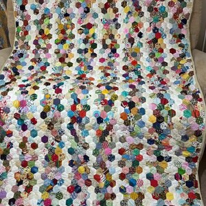 May include: A handmade quilt with a white background and a pattern of colorful, hexagonal patches. The patches are made from various fabrics in a variety of colors and patterns, including floral, stripes, and polka dots. The quilt is draped over a chair.