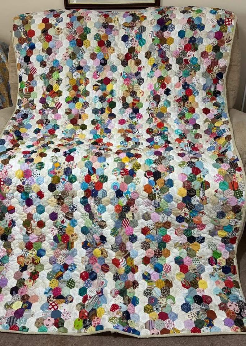 May include: A handmade quilt with a white background and a pattern of colorful, hexagonal patches. The patches are made from various fabrics in a variety of colors and patterns, including floral, stripes, and polka dots. The quilt is draped over a chair.
