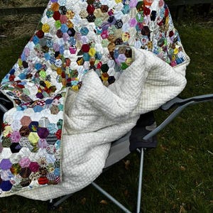 May include: A colorful patchwork quilt with a hexagonal pattern, draped over a folding chair. The quilt features a variety of fabric patterns and colors, including red, blue, yellow, and green. A white, textured blanket is also on the chair.