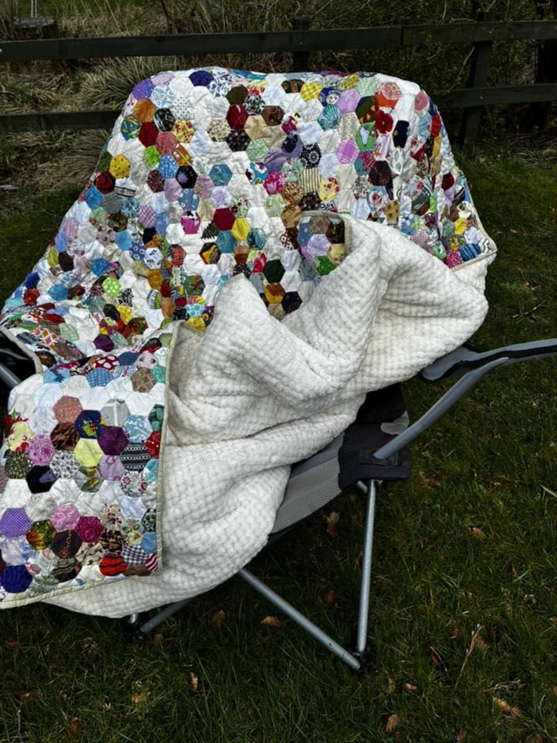 May include: A colorful patchwork quilt with a hexagonal pattern, draped over a folding chair. The quilt features a variety of fabric patterns and colors, including red, blue, yellow, and green. A white, textured blanket is also on the chair.
