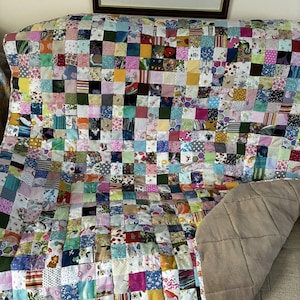 May include: A colourful patchwork quilt made from small, square fabric pieces in a variety of patterns and hues. The quilt is draped over a surface, showing a tan backing. The design includes floral, geometric, and abstract patterns.