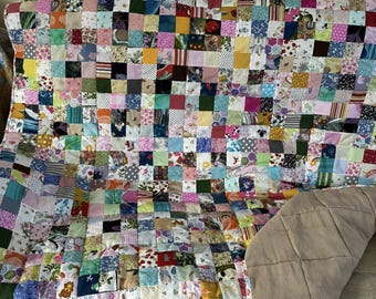 May include: A patchwork quilt with a vibrant mix of small, square fabric pieces in various colors and patterns. The quilt is draped over a surface, revealing a tan backing. The quilt's design includes floral, geometric, and abstract patterns.