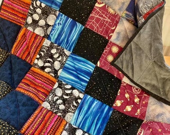 May include: A patchwork quilt with a space theme. The quilt is made of various fabrics with different patterns, including stars, planets, moons, and galaxies. The quilt is in shades of blue, black, red, and white.