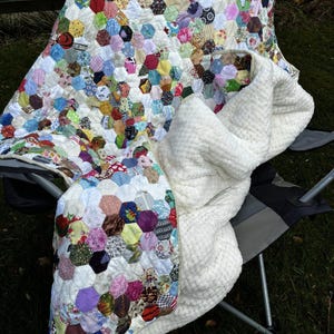 May include: A handmade quilt with a patchwork design of colorful hexagonal fabric pieces. The quilt is draped over a chair, revealing a soft, white, textured blanket underneath. The quilt's colors include red, blue, green, yellow, and purple.