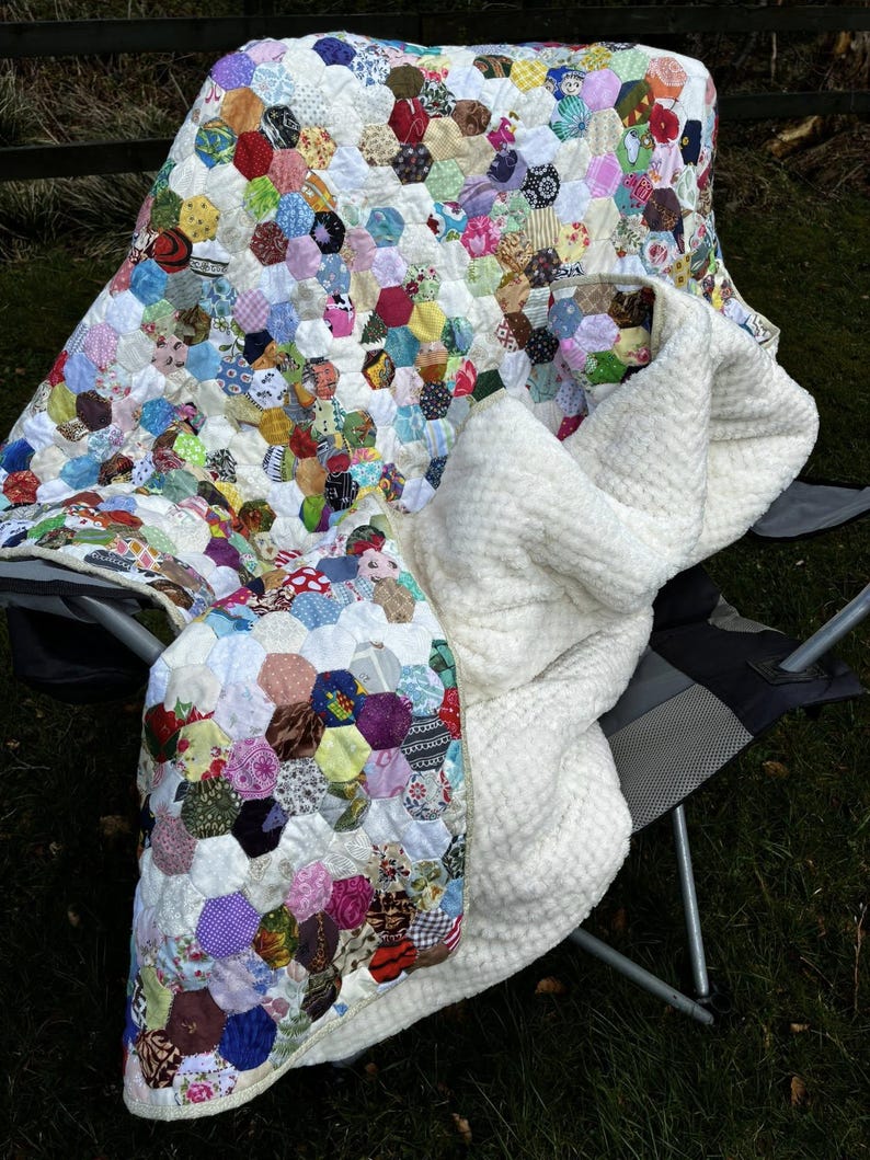 May include: A handmade quilt with a patchwork design of colorful hexagonal fabric pieces. The quilt is draped over a chair, revealing a soft, white, textured blanket underneath. The quilt's colors include red, blue, green, yellow, and purple.