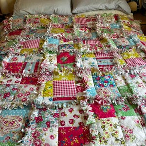 May include: A handmade rag quilt featuring a patchwork design of various floral, gingham, and polka dot fabric squares in shades of pink, red, green, yellow, and blue. The quilt has exposed, frayed seams between each square, creating a textured border.