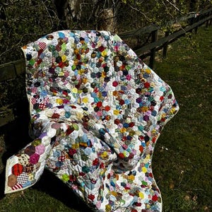 May include: A handmade quilt with a white background and a pattern of colorful, irregularly shaped patches. The quilt is draped over a wooden fence and green grass. The patches are in various colors and patterns, including red, blue, yellow, and green.