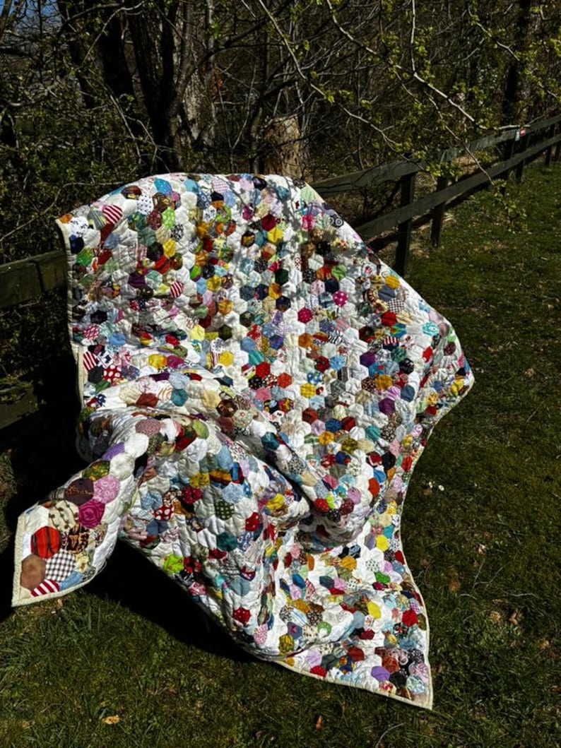 May include: A handmade quilt with a white background and a pattern of colorful, irregularly shaped patches. The quilt is draped over a wooden fence and green grass. The patches are in various colors and patterns, including red, blue, yellow, and green.