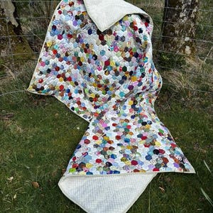 May include: A colorful patchwork quilt with a white backing, displayed outdoors. The quilt features a variety of patterned hexagonal fabric pieces in shades of red, blue, green, and yellow. The quilt is draped over a wire fence and grass.