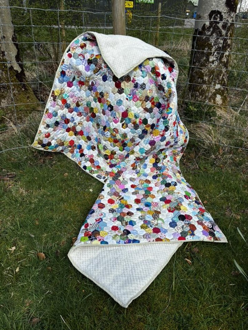 May include: A colorful patchwork quilt with a white backing, displayed outdoors. The quilt features a variety of patterned hexagonal fabric pieces in shades of red, blue, green, and yellow. The quilt is draped over a wire fence and grass.