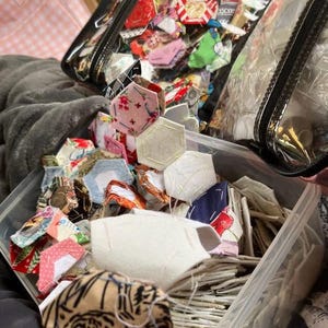 May include: A clear plastic container filled with colorful fabric pieces, including hexagons and other shapes. The fabrics feature various patterns and colors, such as floral prints, stripes, and solid colors. A clear zippered bag is also visible, filled with similar fabric pieces.