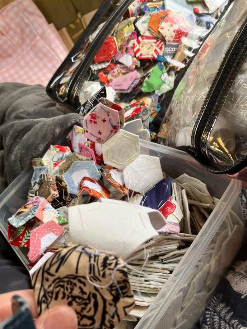 May include: A clear plastic container filled with colorful fabric pieces, including hexagons and other shapes. The fabrics feature various patterns and colors, such as floral prints, stripes, and solid colors. A clear zippered bag is also visible, filled with similar fabric pieces.