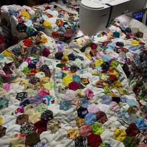 May include: A handmade quilt in progress, featuring a patchwork design of colorful, circular fabric pieces sewn onto a white background. A sewing machine is positioned on the quilt, with the needle poised to stitch the fabric. The quilt is covered in a variety of colors and patterns.