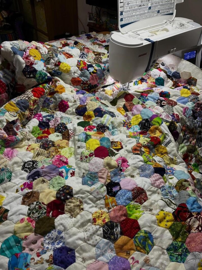 May include: A handmade quilt in progress, featuring a patchwork design of colorful, circular fabric pieces sewn onto a white background. A sewing machine is positioned on the quilt, with the needle poised to stitch the fabric. The quilt is covered in a variety of colors and patterns.