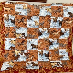 May include: A quilted throw blanket with a patchwork design. It features squares of running horses against brown and blue backgrounds. The blanket has a red border with a geometric pattern and is draped over a beige armchair.