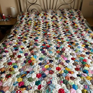 May include: A handmade quilt with a colorful patchwork design. The quilt features a white background with numerous hexagonal patches in various colors and patterns, including red, blue, green, and yellow. The quilt is spread across a bed with a decorative metal headboard.