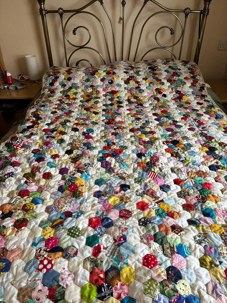 May include: A handmade quilt with a colorful patchwork design. The quilt features a white background with numerous hexagonal patches in various colors and patterns, including red, blue, green, and yellow. The quilt is spread across a bed with a decorative metal headboard.
