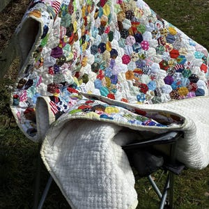 May include: A handmade quilt with a patchwork design of colorful fabric hexagons. The quilt is draped over a folding chair, revealing a soft, white, quilted backing. The quilt's vibrant colors include red, blue, yellow, and green.