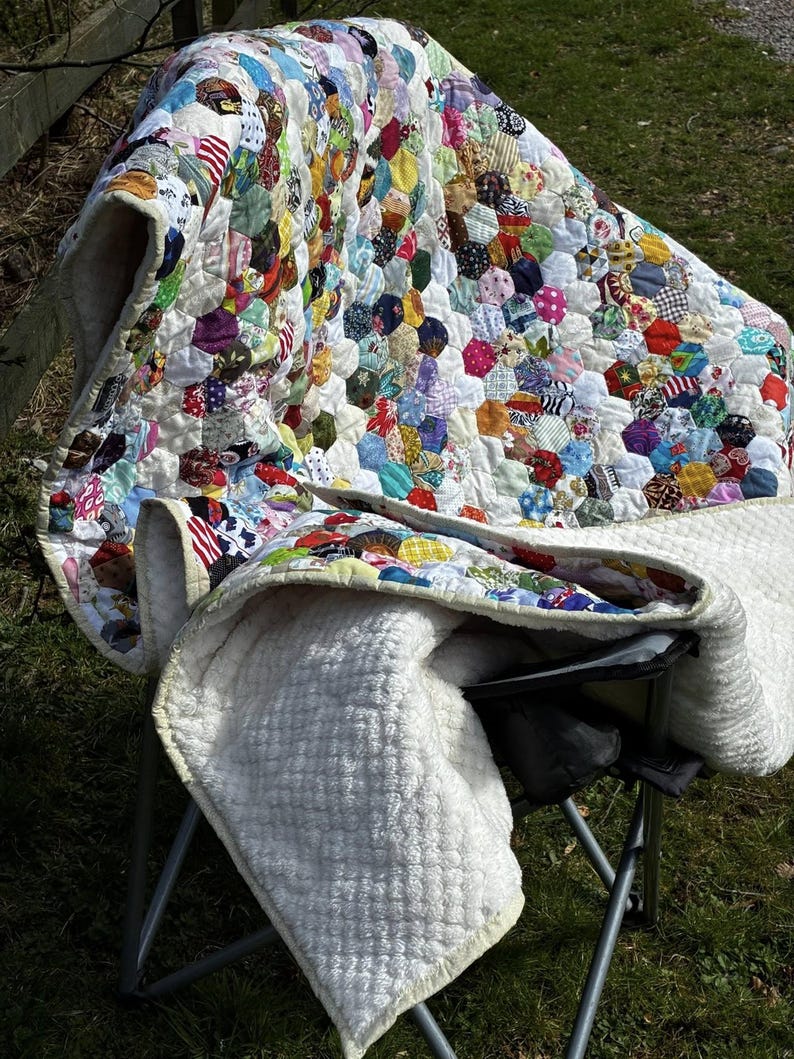 May include: A handmade quilt with a patchwork design of colorful fabric hexagons. The quilt is draped over a folding chair, revealing a soft, white, quilted backing. The quilt's vibrant colors include red, blue, yellow, and green.