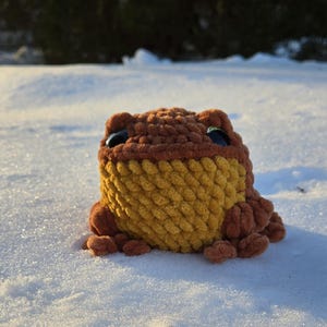 May include: A handmade crochet frog toy in shades of brown and yellow, with large black eyes. The frog is sitting in the snow, showcasing its textured, plush design. The toy is a whimsical, handcrafted item.