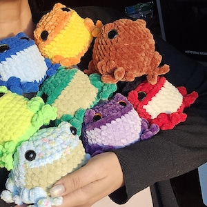 May include: A collection of handmade crochet frog plushies in various colors, including blue, orange, brown, green, purple, and red. Each frog has large black eyes and is made with a soft, textured yarn. The frogs are a whimsical and colorful assortment.