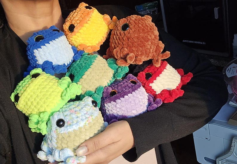 May include: A collection of handmade crochet frog plushies in various colors, including blue, orange, brown, green, purple, and red. Each frog has large black eyes and is made with a soft, textured yarn. The frogs are a whimsical and colorful assortment.