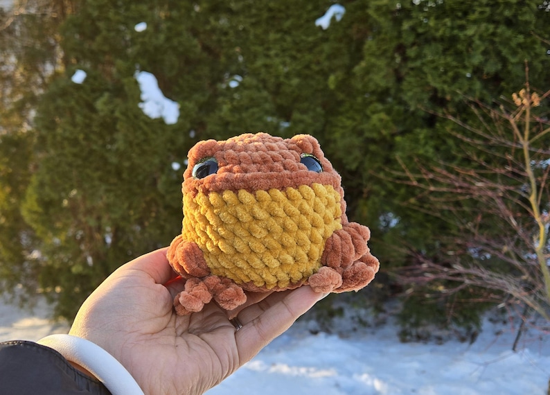 May include: A handmade crochet frog in shades of brown and yellow, with large blue eyes. The plush toy is held in a hand, with a blurred background of green foliage and sunlight.