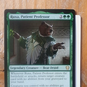 May include: A green, black and white Magic: The Gathering card featuring a bear druid named Ruxa, Patient Professor. The card text reads: "Whenever Ruxa, Patient Professor enters the battlefield or attacks, return target creature card with no abilities from your graveyard to your hand. Creatures you control with no abilities get +1/+1. You may have creatures you control with no abilities assign their combat damage as though they weren't blocked."