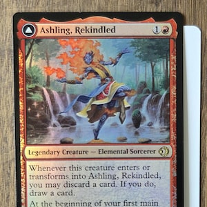May include: A Magic: The Gathering card featuring "Ashling, Rekindled." The card has a red and black border with an illustration of a blue-skinned figure with fiery hair. Text includes "Legendary Creature - Elemental Sorcerer" and game instructions. The card is 1/3.