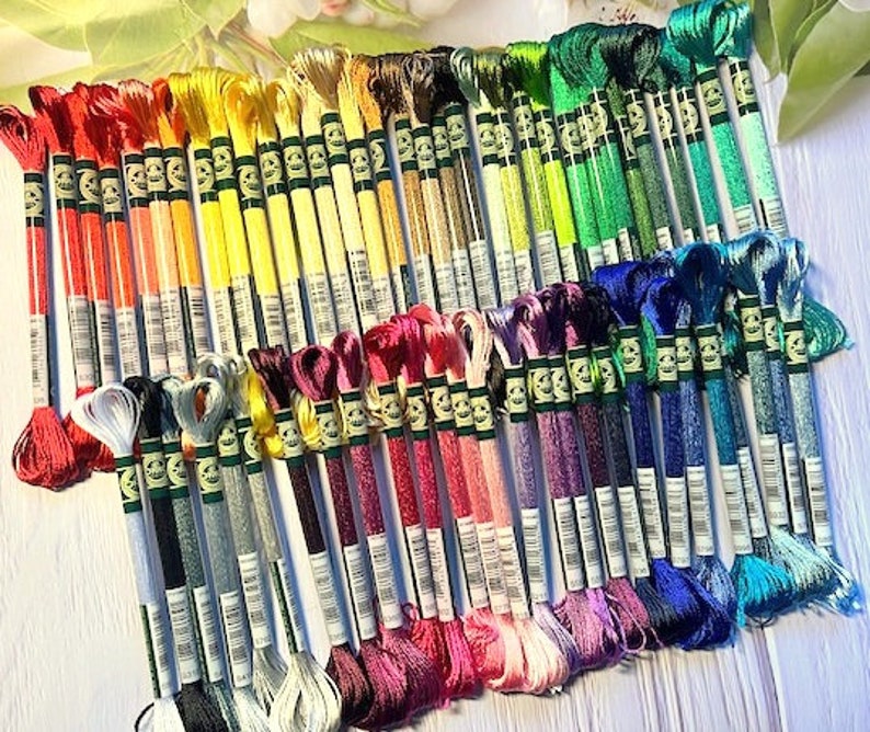 Puede incluir: Una colecci&oacute;n de carretes de hilo de bordar en un arco&iacute;ris de colores, dispuestos en filas. Los carretes est&aacute;n etiquetados con n&uacute;meros y tienen una etiqueta verde y blanca.