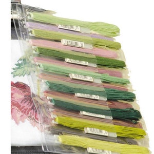 May include: A clear plastic organiser sheet containing multiple skeins of embroidery floss in various shades of green, pink, and purple. Each skein is attached to a white paper tag with a printed barcode, arranged in a vertical row.