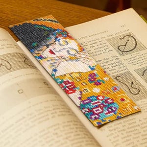 May include: A cross-stitch bookmark, inspired by Gustav Klimt's "The Kiss", with a colourful design in gold, blue, red, and black. The bookmark is placed in an open book with text and illustrations, perfect for book lovers.