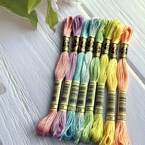 May include: Eight spools of DMC embroidery floss in pastel colors, including pink, lavender, light blue, green, yellow, and peach. Each spool has a black label with the DMC logo and a number printed on it. The numbers are 354, 554, 3766, 828, 164, 3819, 744, and 3824.