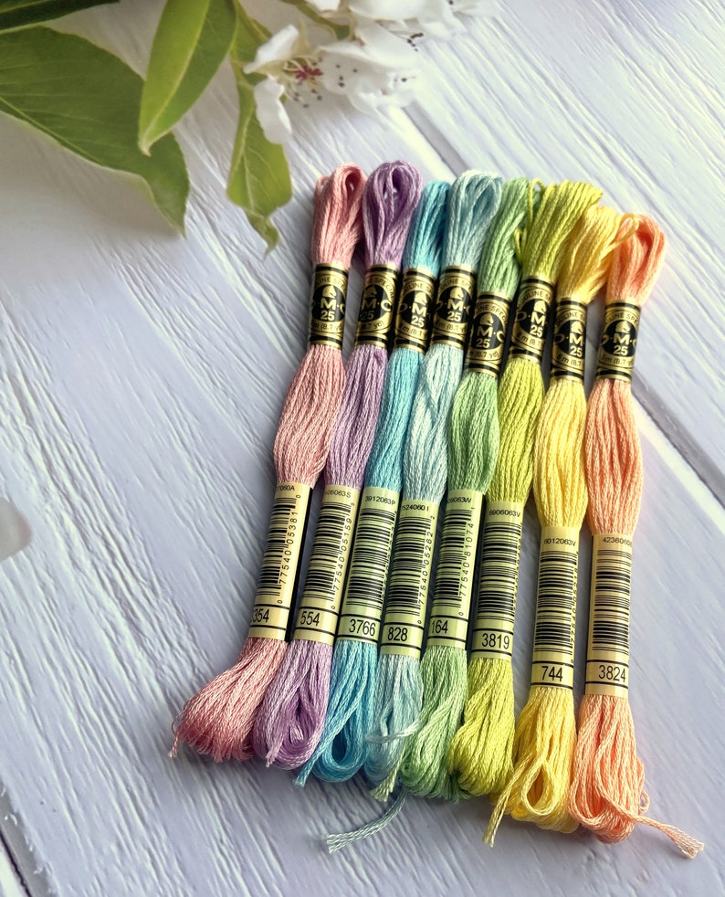 May include: Eight spools of DMC embroidery floss in pastel colors, including pink, lavender, light blue, green, yellow, and peach. Each spool has a black label with the DMC logo and a number printed on it. The numbers are 354, 554, 3766, 828, 164, 3819, 744, and 3824.