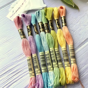 May include: Eight skeins of DMC embroidery floss in pastel colors, including pink, lavender, light blue, green, and yellow. Each skein has a DMC label with a barcode and the DMC number printed on it.