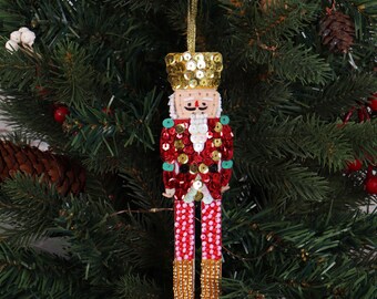Peut inclure: Une décoration casse-noisette ornée d'un chapeau à sequins dorés, d'une veste à sequins rouges avec des accents verts et d'une barbe en perles blanches. Les jambes sont décorées de perles roses et rouges, et les pieds de perles dorées, suspendus.