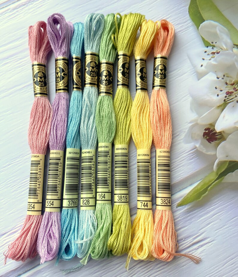 May include: Eight spools of DMC embroidery floss in pastel colors. The colors are: light pink, lavender, light blue, light green, yellow, light orange, and peach. Each spool has a white label with black text and a barcode. The text on the label includes "DMC", "Mouline Special", "25", and a number.