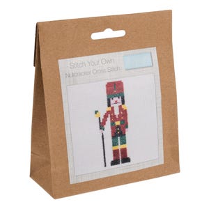 May include: A brown paper bag containing a white card with a red and green cross stitch design of a nutcracker soldier. The text on the card reads "Stitch Your Own Nutcracker Cross Stitch" and "Tonic"