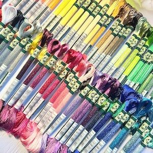 Puede incluir: Una colecci&oacute;n de carretes de hilo de bordar en un arco&iacute;ris de colores, incluyendo blanco, azul claro, rosa, morado, rojo, naranja, amarillo, verde y azul. Los carretes est&aacute;n dispuestos en forma de abanico, con los colores mezcl&aacute;ndose entre s&iacute;.