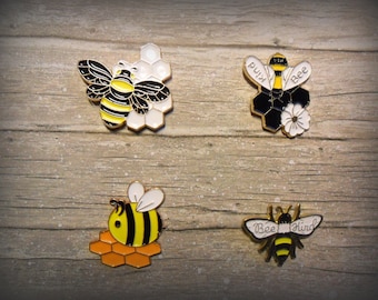 May include: Four enamel pins featuring bees and honeycomb designs. The pins are gold-colored with black and yellow accents. One pin says "Bee Kind", another says "Bee", and the other two feature bees on honeycomb.